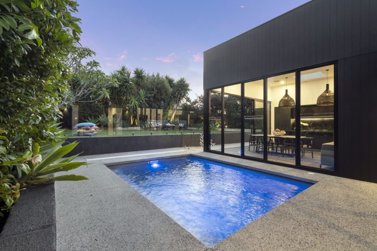 A sleek courtyard pool with tropical landscaping, glass fencing, and a black modern home.